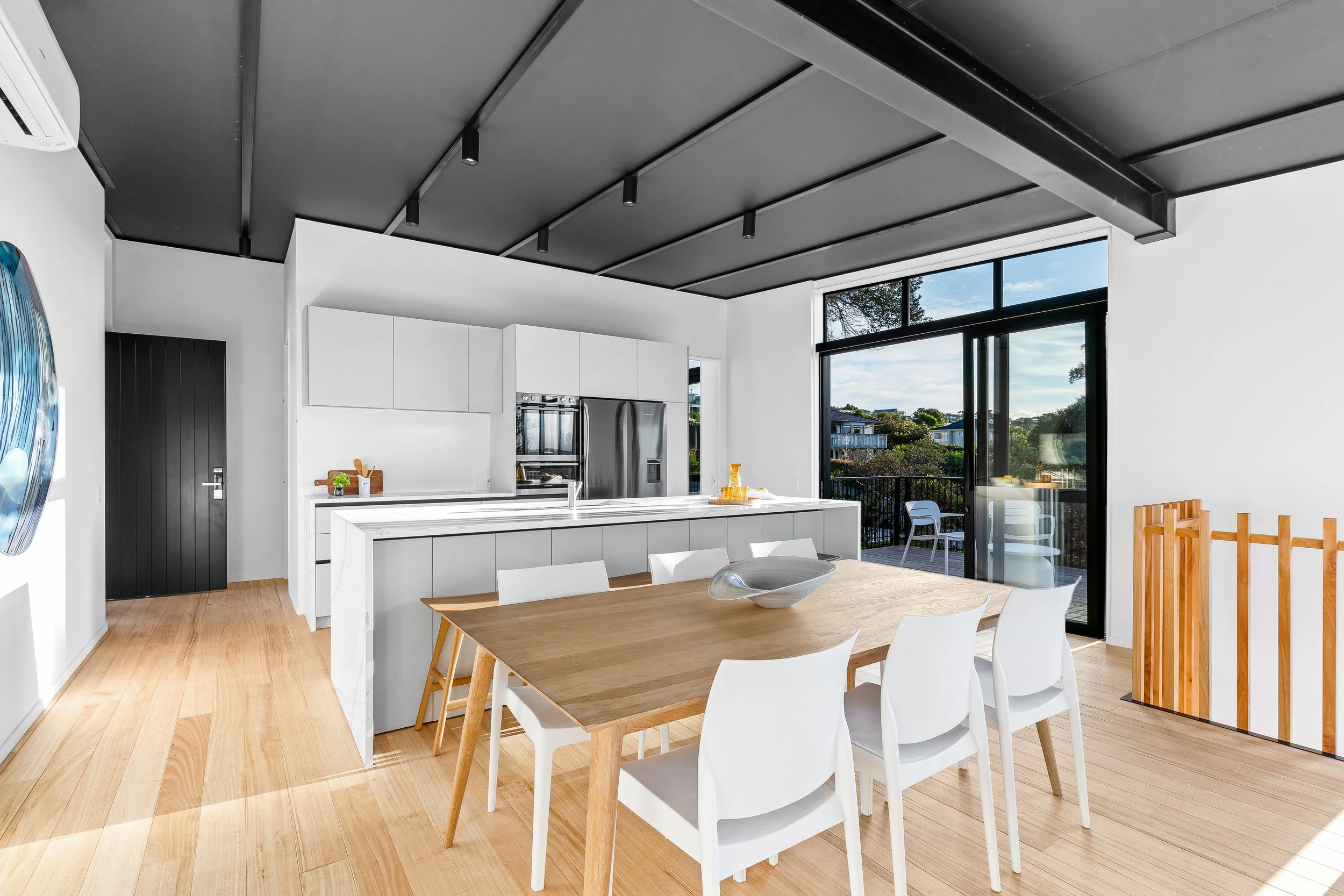 Open-concept kitchen and dining area with white cabinetry, a wooden dining table, white chairs, and large sliding glass doors leading to an outdoor balcony represents new build project by gravity plumbing