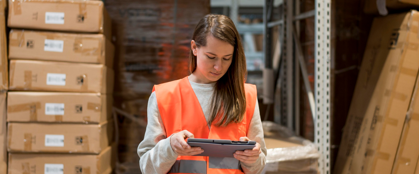 A woman in an orange safety vest standing in a warehouse, looking down at a clipboard.