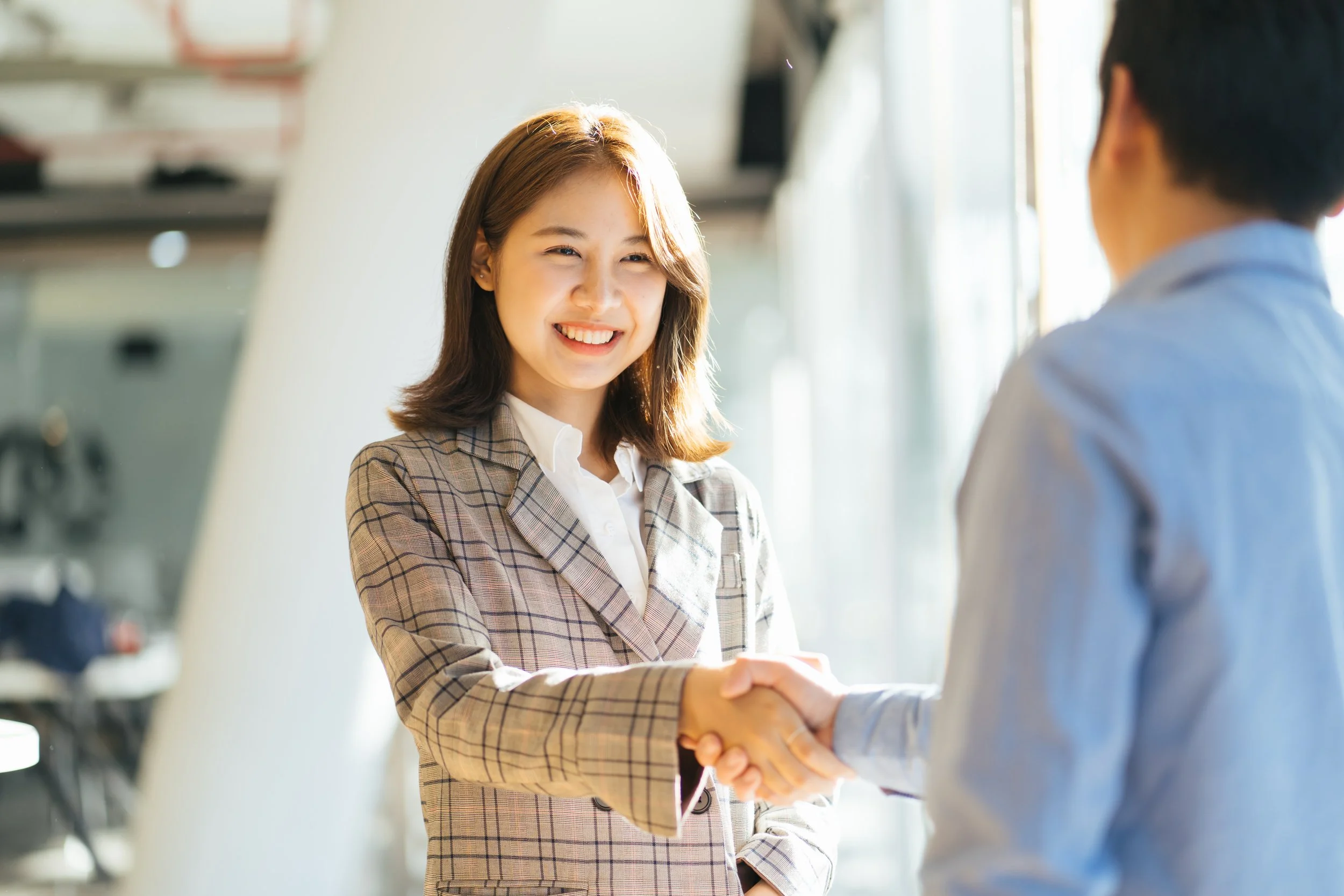 A woman with shoulder-length hair smiling and shaking hands with a person whose face is not visible. She is wearing a plaid blazer and a white shirt, inside a well-lit room with shelves and a window in the background.