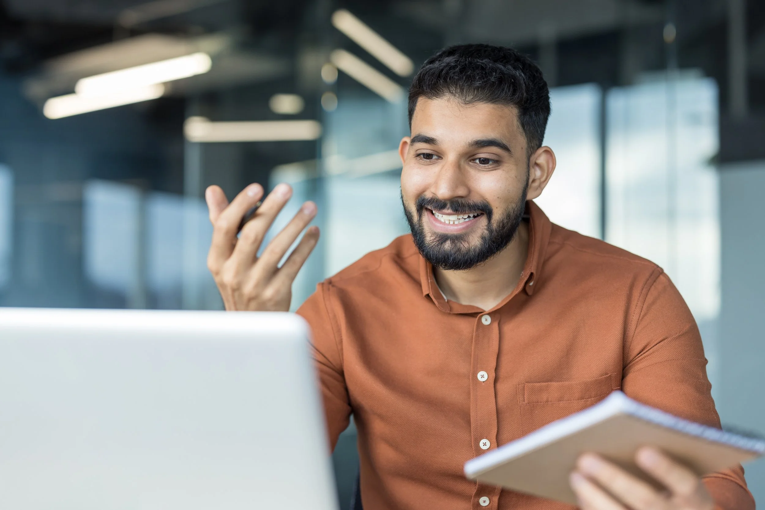 A man with dark hair, a beard, and wearing a brown shirt, smiling and holding a notebook while looking at his phone in a modern office setting.