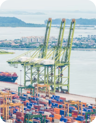 Large container port with stacked shipping containers and three tall green cranes over the water, with a cityscape in the background.