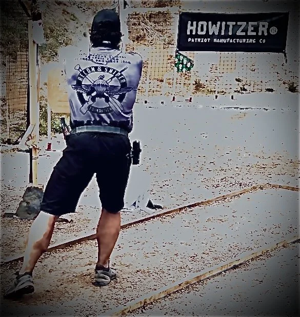 A man shooting a rifle at an indoor shooting range. The man is wearing tactical clothing and safety equipment. There's a sign that reads 'HOWITZER Patriot Manufacturing Co' on the wall.