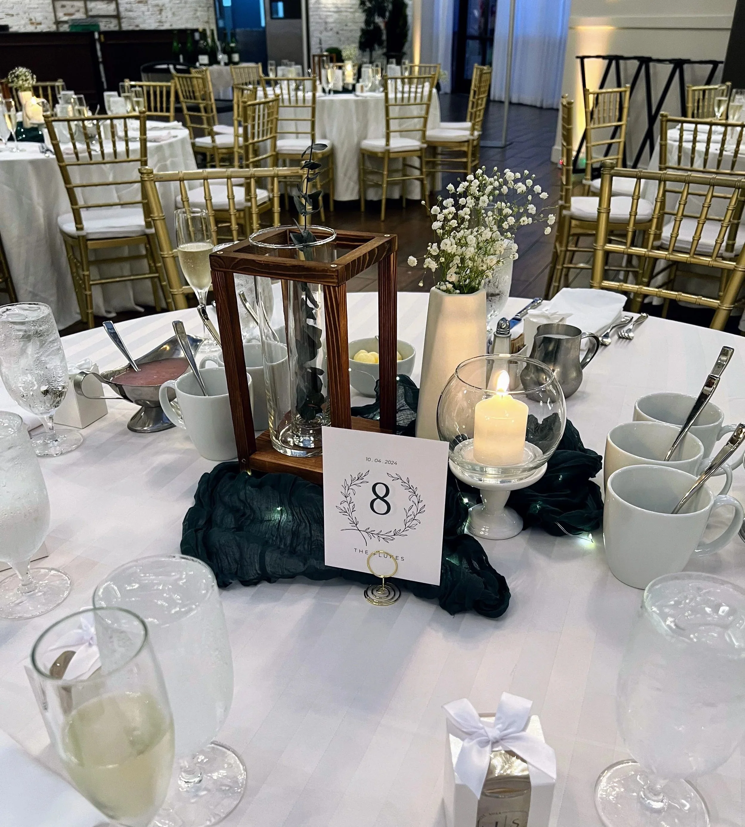 Wedding reception table with gold chairs, white tablecloths, floral centerpieces, candles, and place settings, decorated in an elegant style in a modern venue.