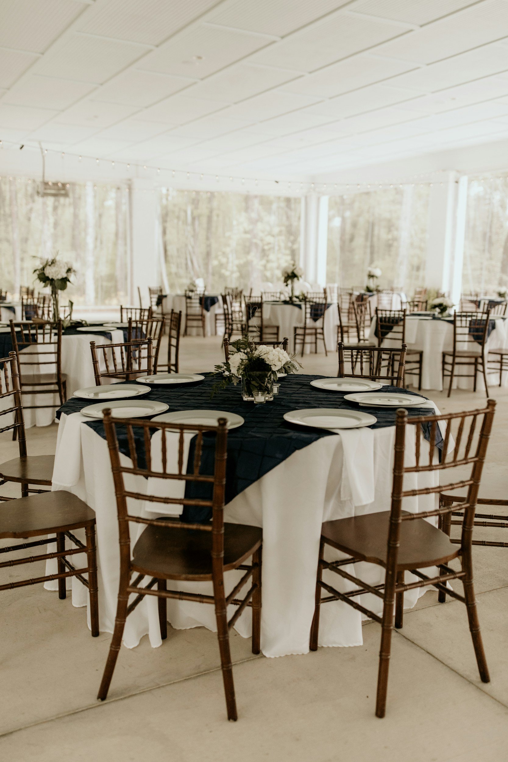 Round banquet tables covered with white tablecloths and black table runners, set with white plates, silverware, and centerpieces of white flowers in vases, in a well-lit, indoor venue with a textured wall and string lights overhead.