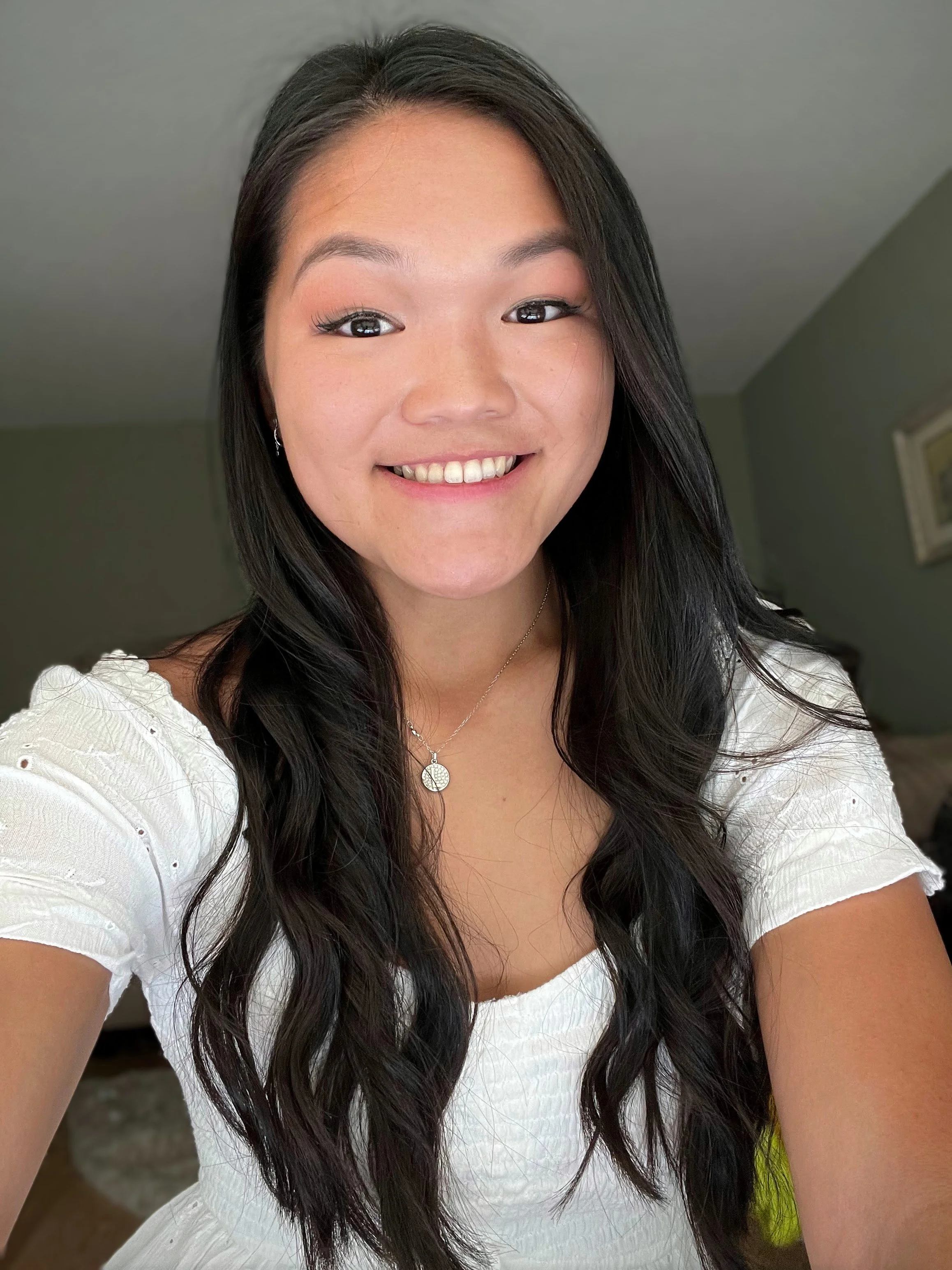 Close-up of a smiling young woman with long dark hair, wearing a white top and jewelry, in a room with green walls.