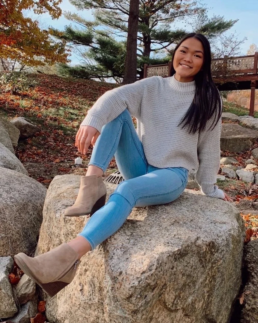 A young woman with long dark hair smiling and seated on a large rock outdoors in autumn, wearing a light gray sweater, light blue jeans, and beige ankle boots, surrounded by fall foliage and trees.