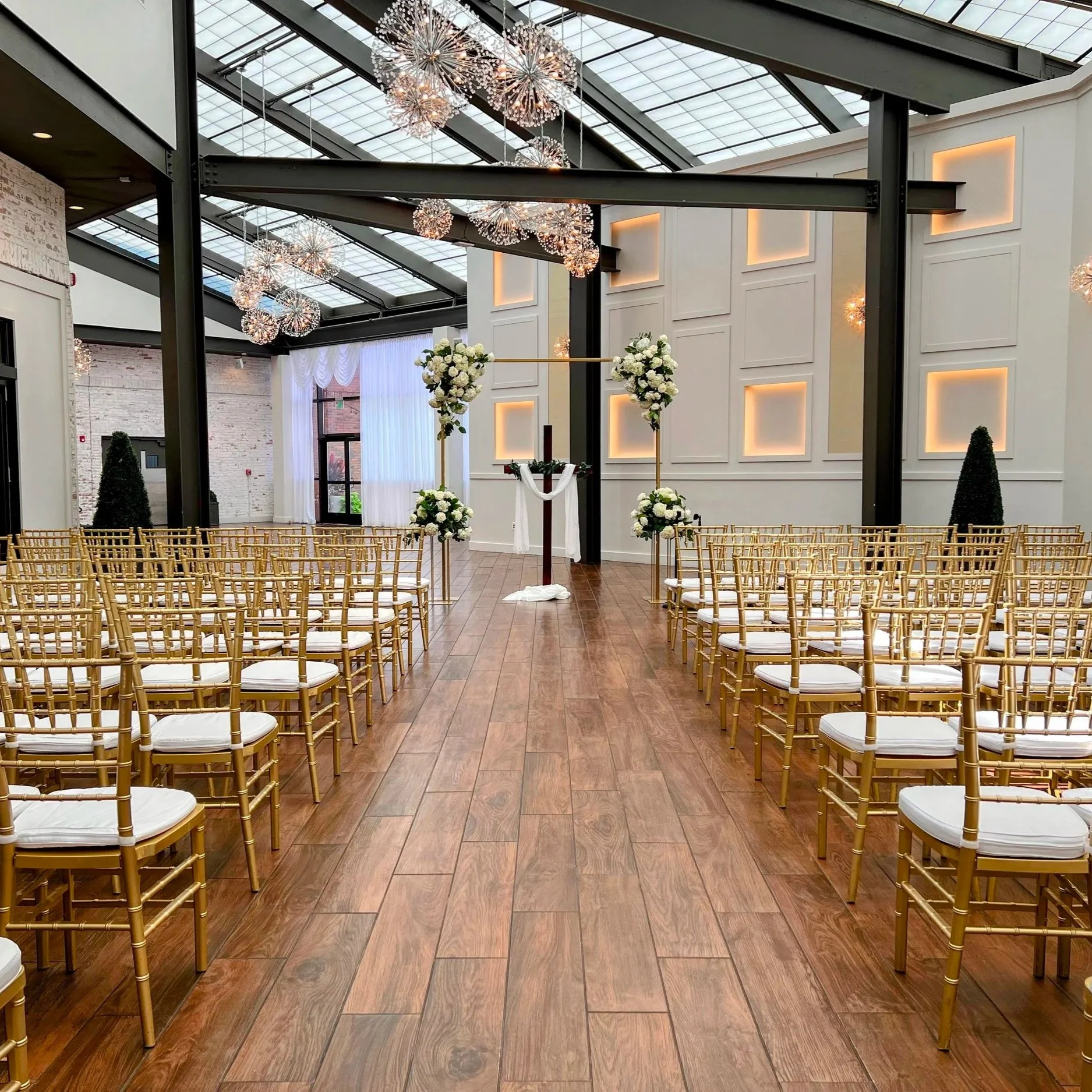Interior of a modern event space decorated for a wedding, with rows of gold chairs, floral arrangements on a cross-shaped stand, hanging spherical light fixtures, a white curtain, and a brick wall in the background.