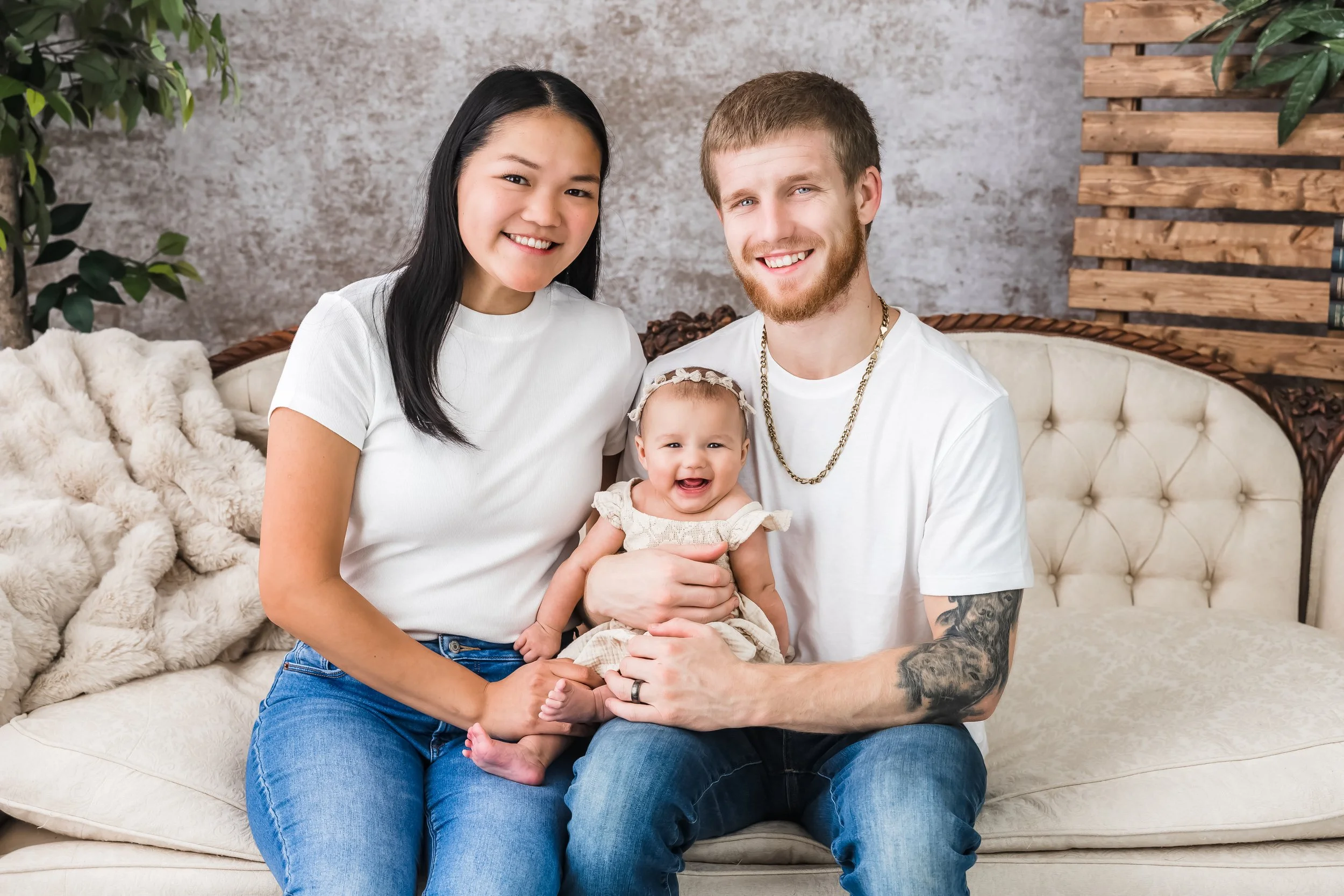 A smiling family of three sitting on a beige couch, including a woman with long black hair, a man with short brown hair and a beard, and a baby girl with light brown hair and a headband, in a living room with a textured wall and wooden accents.