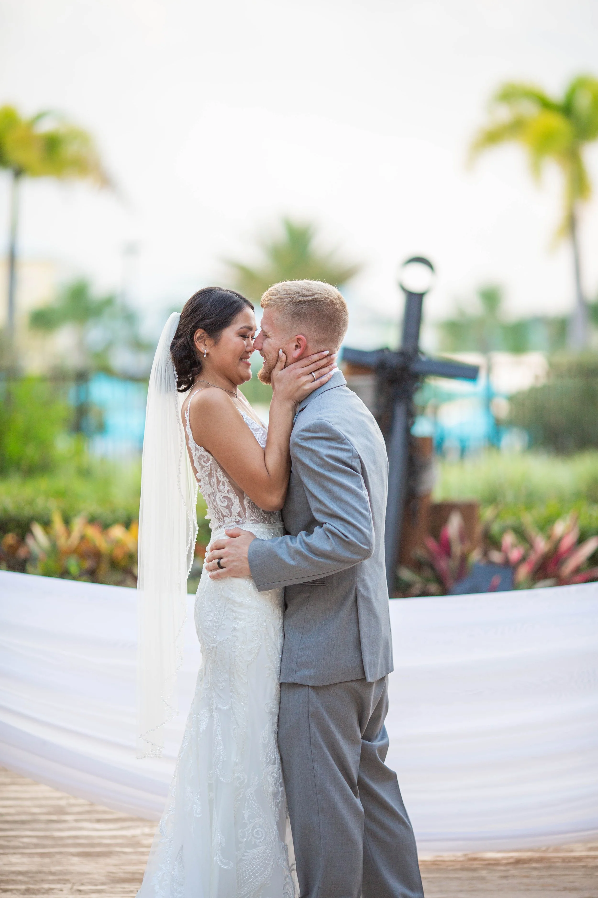 A bride and groom kiss at their wedding ceremony outdoors, with palm trees and colorful plants in the background.