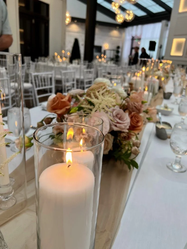 A candle in a glass holder on a decorated banquet table at an event space with floral arrangements, candles, and elegant decor.