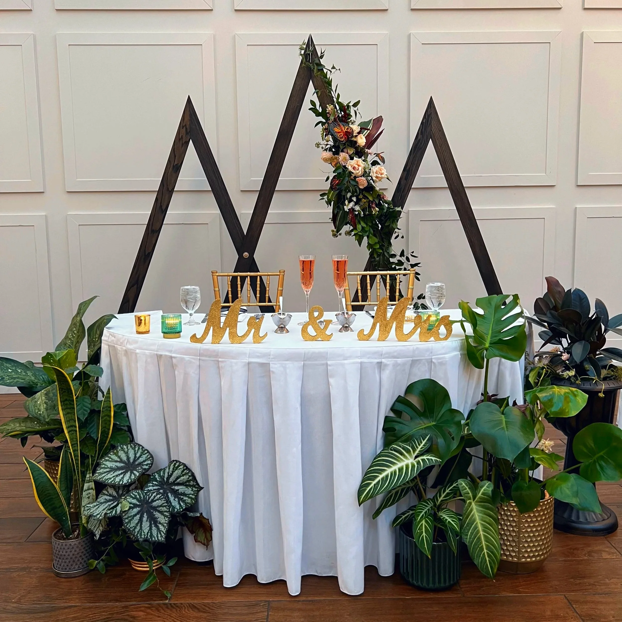 A decorated table with floral arrangements, candles, and the words 'Mr. & Mrs.' in gold on a white tablecloth. The background features a white wall with wood paneling and a black wooden geometric structure with flowers.