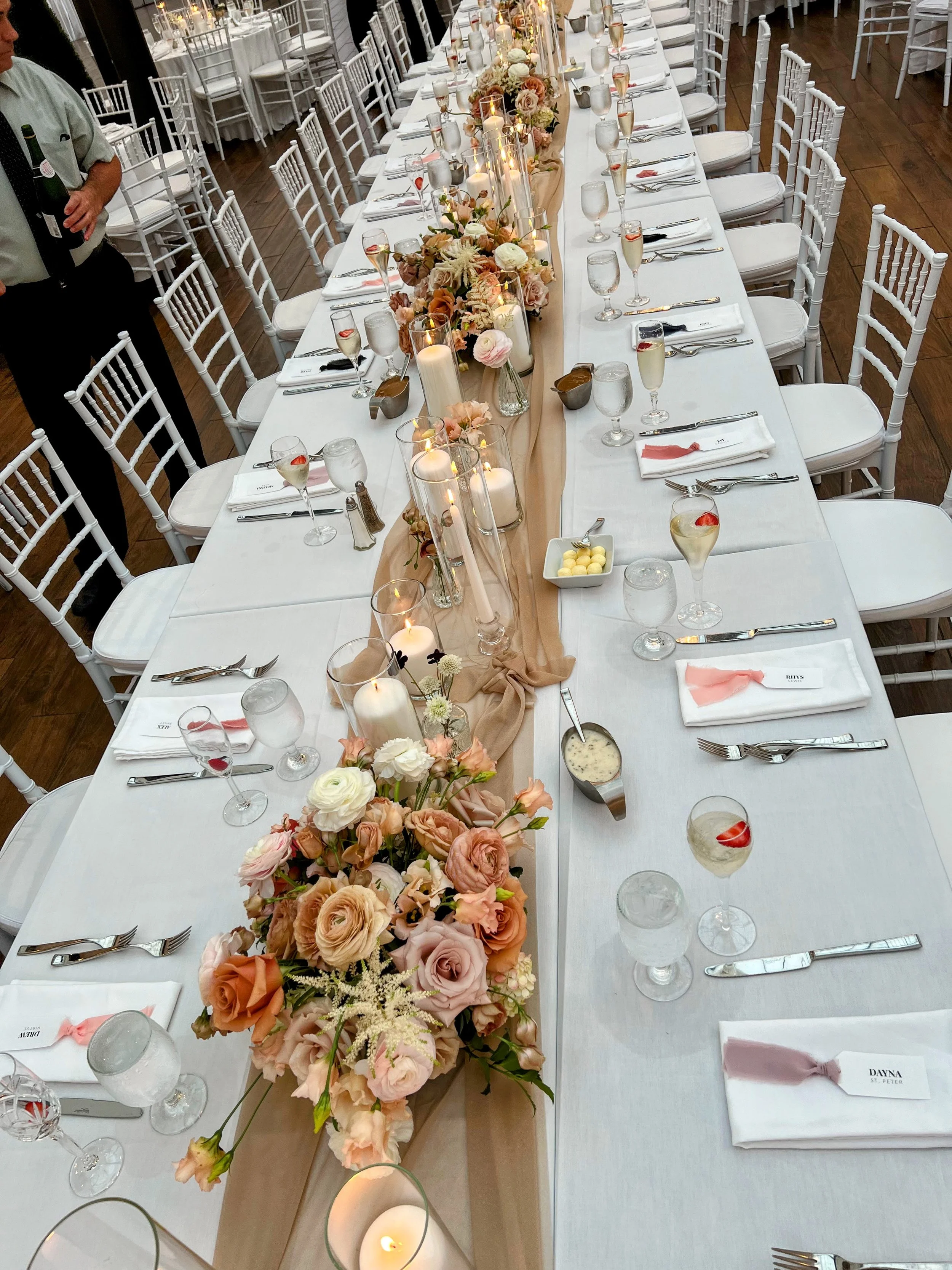 A long banquet table decorated for a wedding reception with floral arrangements, candles, and glasses of champagne. The table is set with white linens, silverware, and cloth napkins, with white chairs arranged along each side.