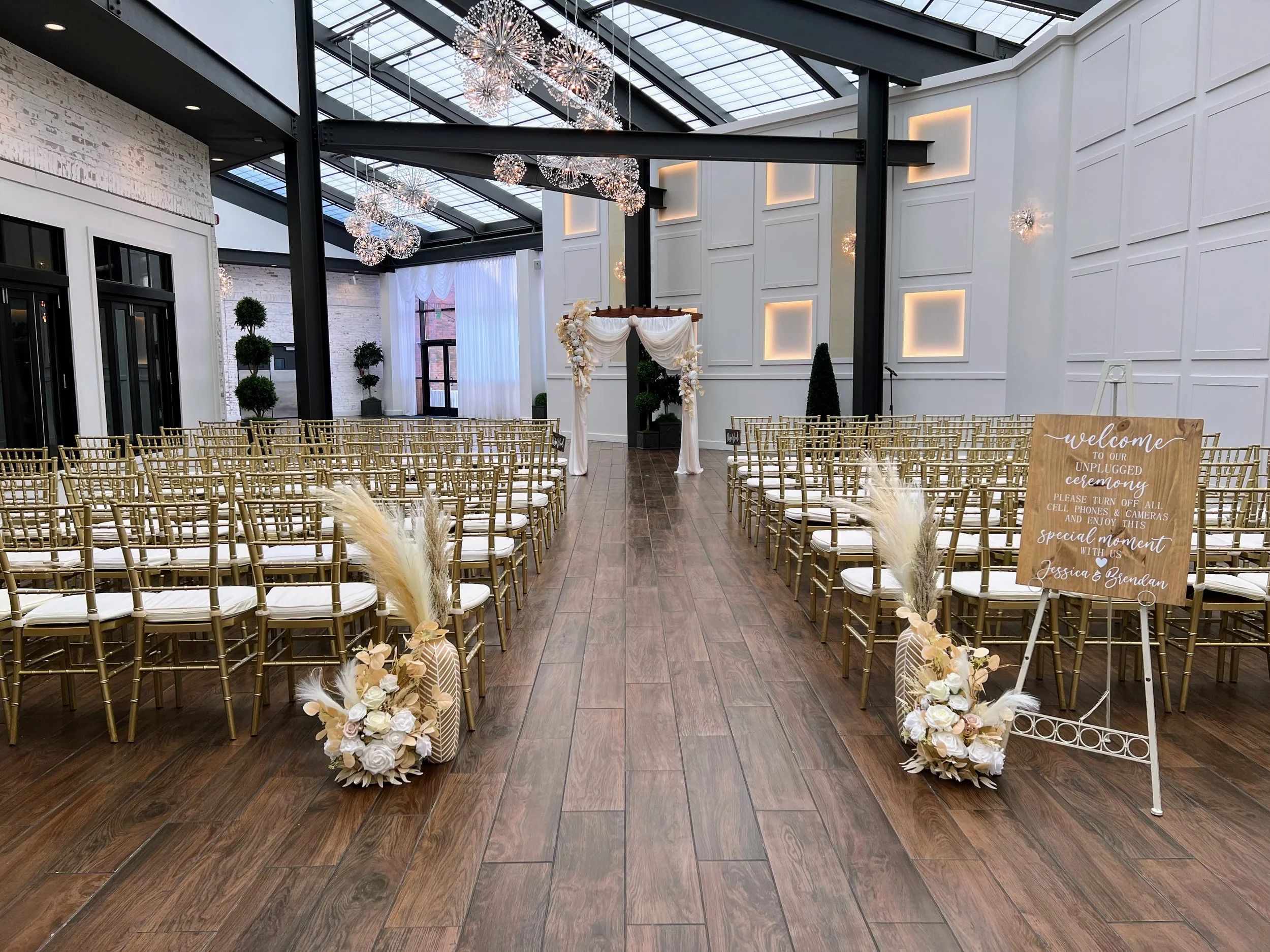Interior of a wedding venue with rows of gold chairs, floral arrangements, and a decorated arch at the front for ceremony. Modern lighting fixtures and a glass roof illuminate the space.