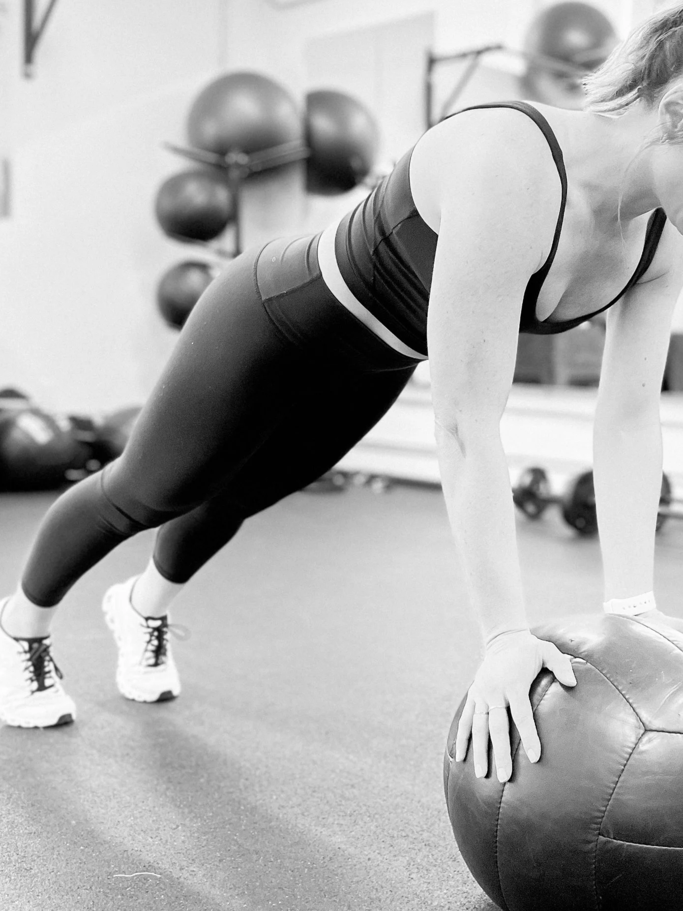 A woman in workout clothes performing a push-up exercise with her hands on a medicine ball, in a gym setting with medicine balls on a rack in the background.