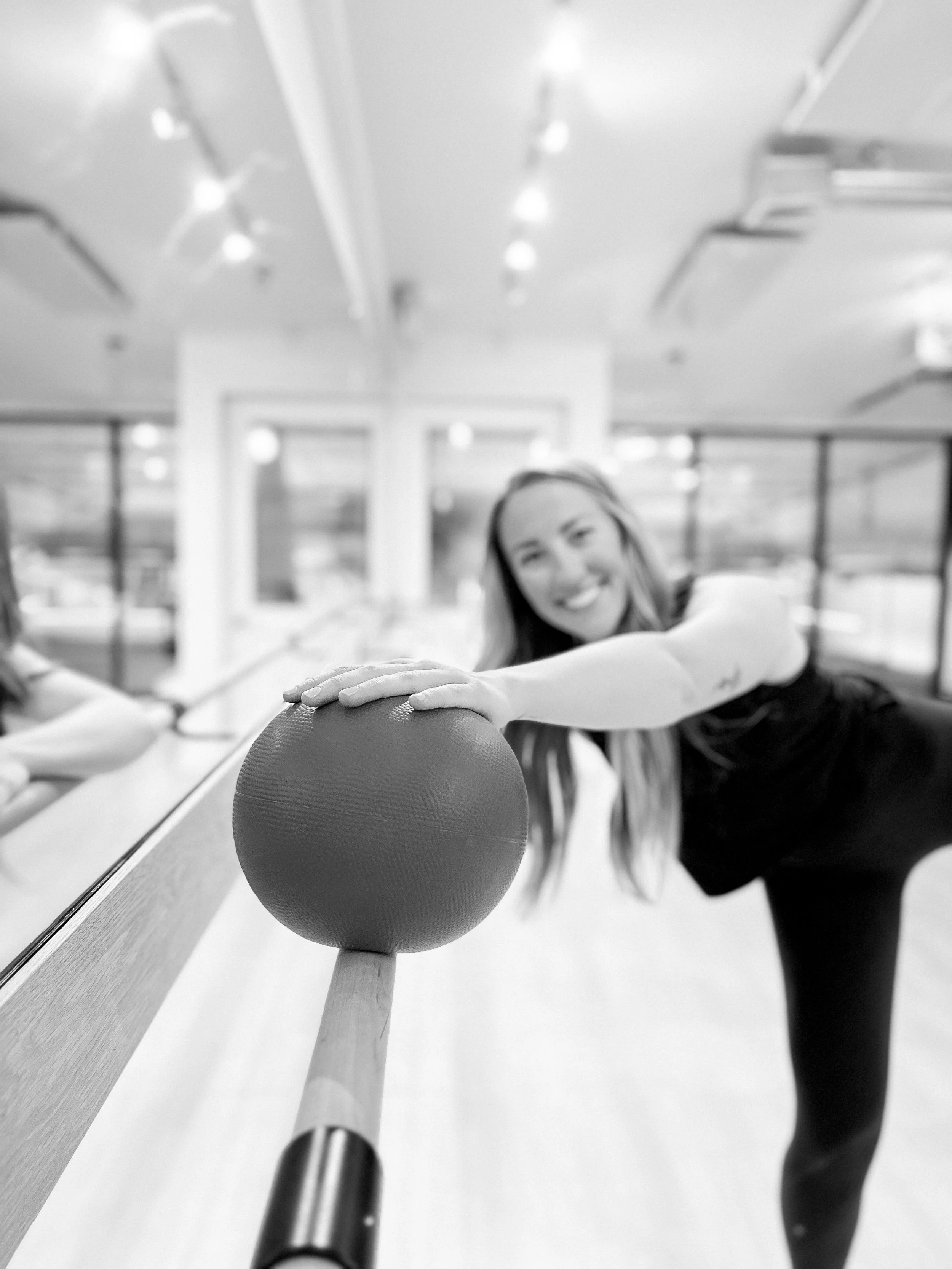 A woman smiling and reaching out her hand to touch a ping pong paddle on a table in a gym or sports room, with a rack of tennis balls and mirrors in the background.