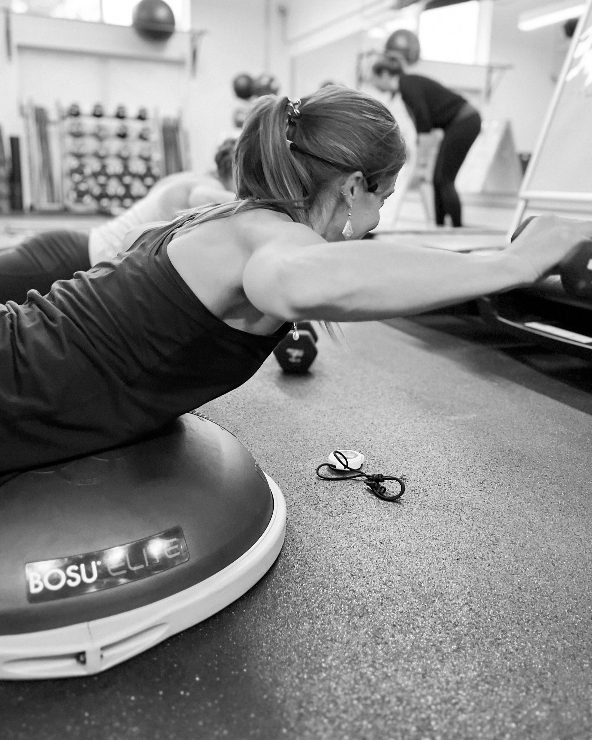 A woman exercising on a mat in a gym, with gym equipment and a person in the background. The photo is in black and white.