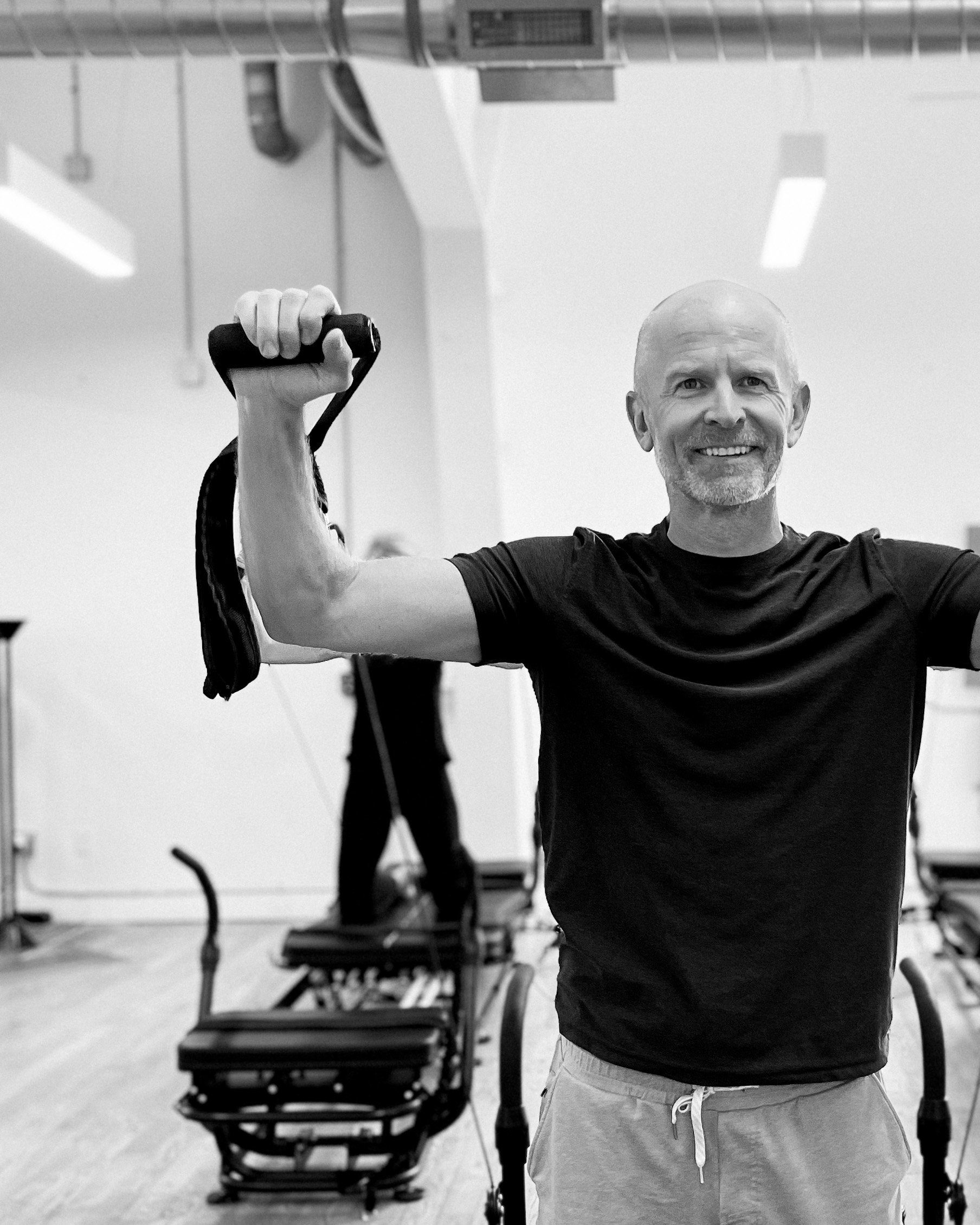 A smiling man flexing his arm at a gym, with exercise equipment in the background.