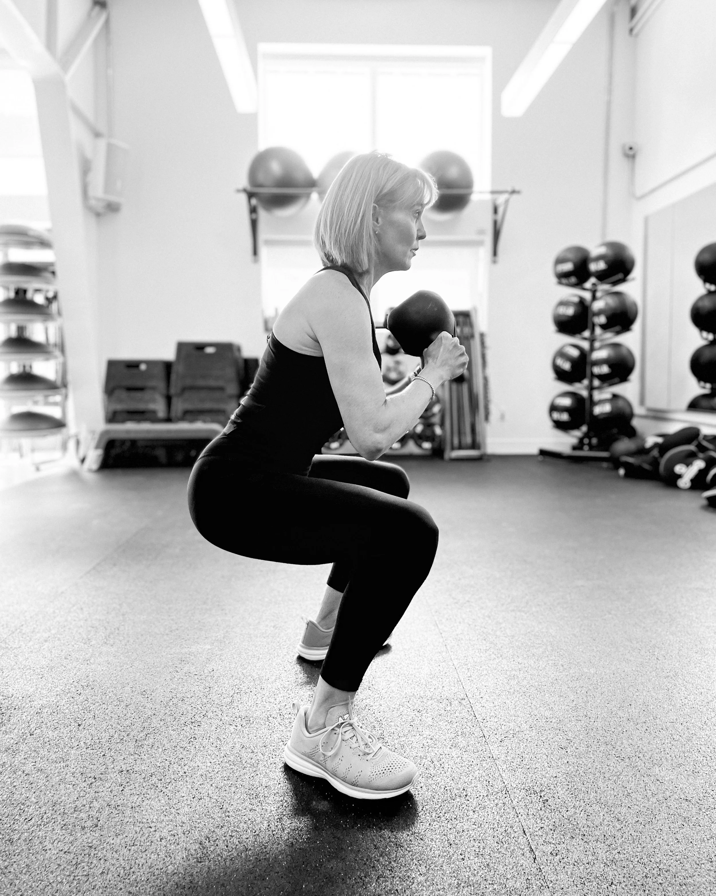 A woman in workout clothes performing a squat with a kettlebell in a gym. She is wearing athletic shoes and has her hair in a bob cut. The gym has shelves with exercise balls and equipment in the background.