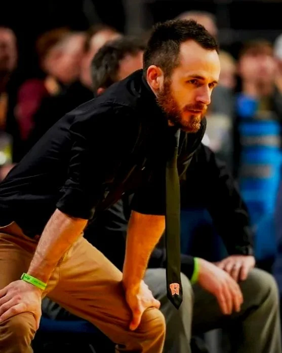 A man with a beard sitting forward, leaning on his hands, at an event with several people in the background.