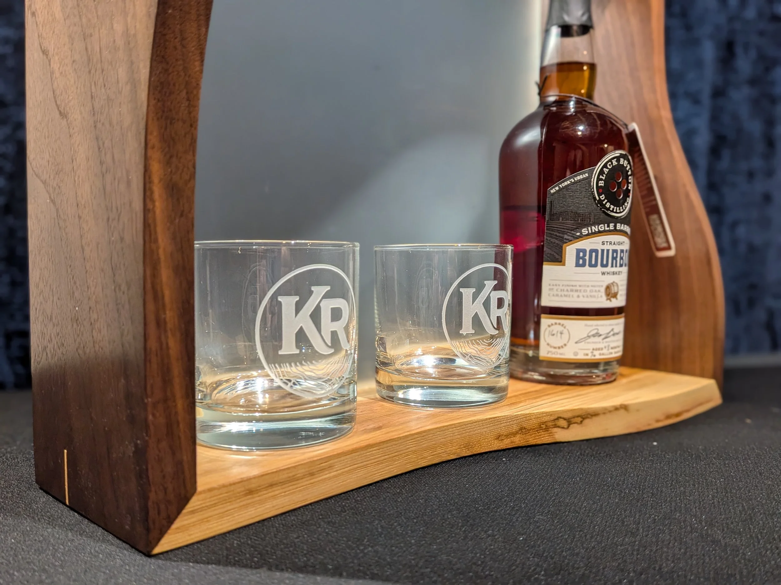 A wooden display shelf holding two glasses and a bottle of bourbon whiskey. The glasses have a logo with the letters 'KR' on them. The whisky bottle is labeled 'Black Buck Bourbon' and has a dark brown liquid inside.