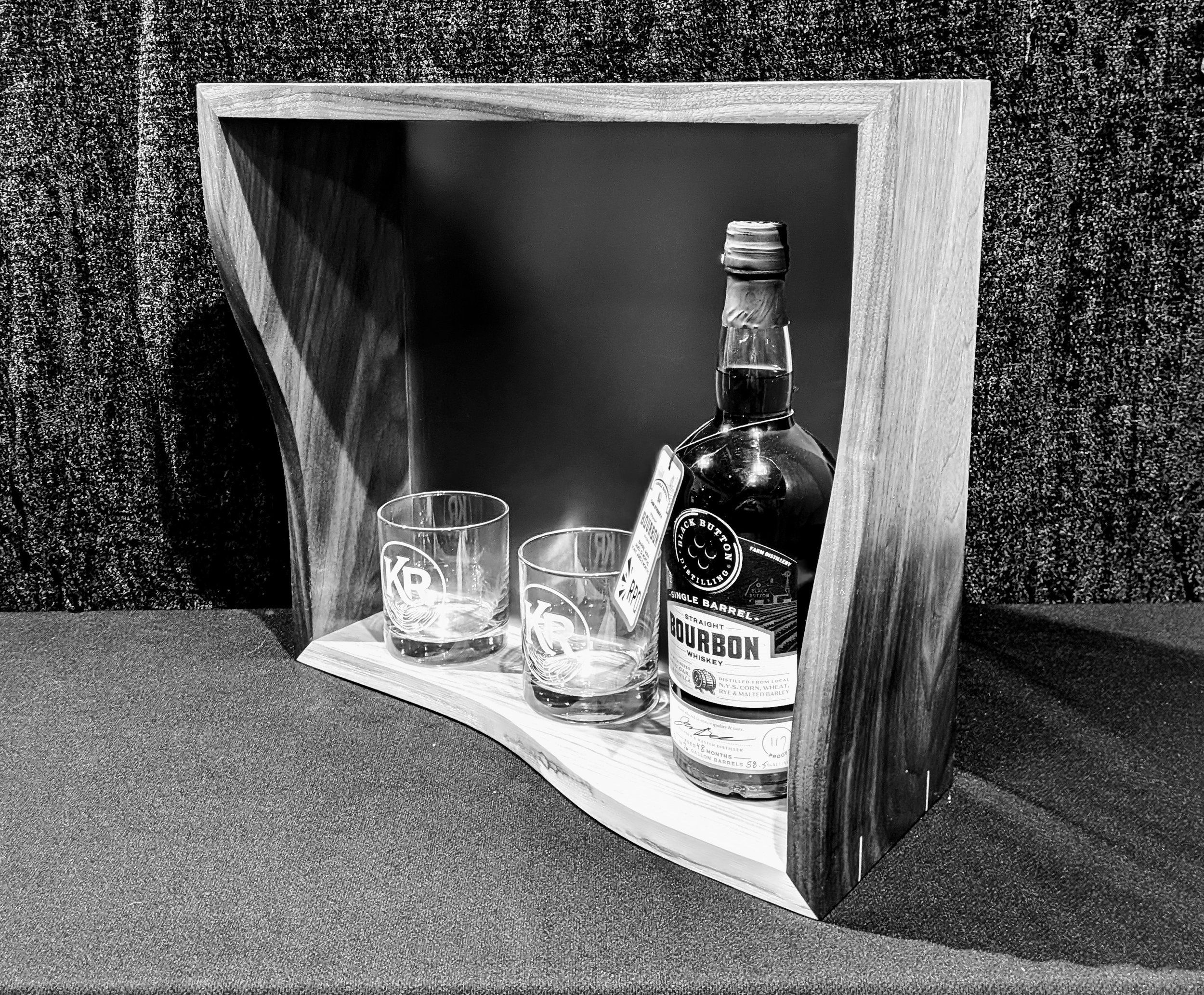 A bottle of whiskey and two empty glasses in front of a wooden picture frame, placed on a textured surface.