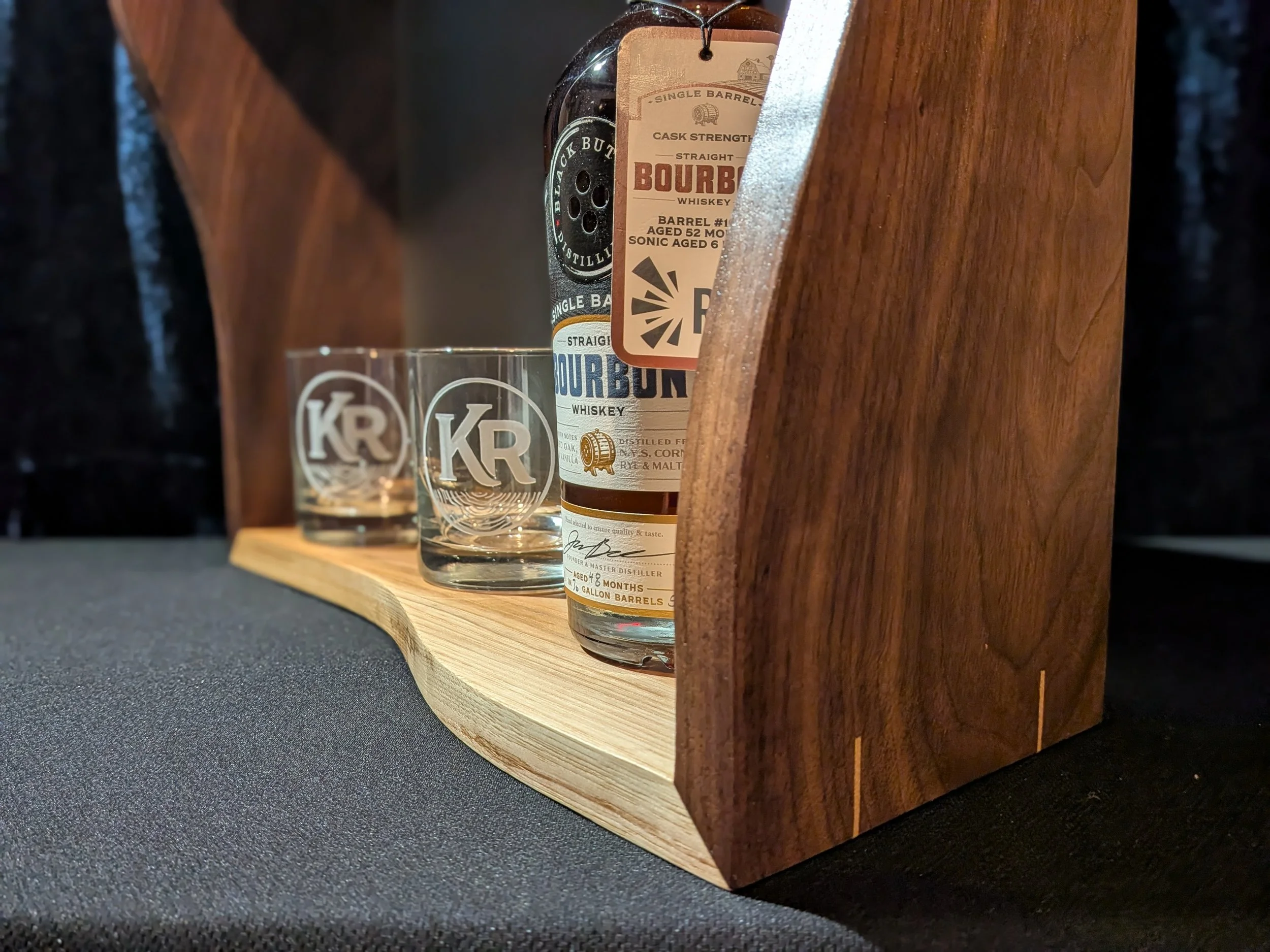 A bottle of bourbon whiskey on a wooden display stand with two whiskey glasses in front with logos, set against a dark background.