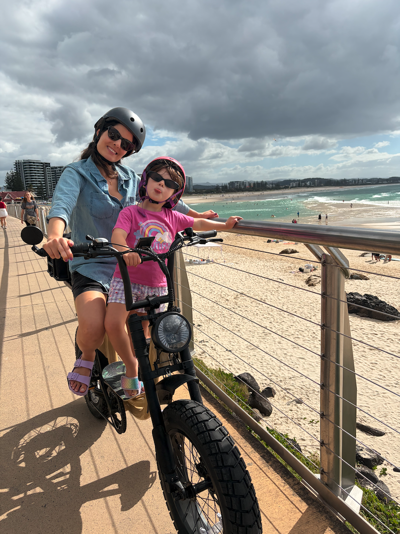 Two girls, wearing helmets and sunglasses, riding a bicycle on a boardwalk by the beach with cloudy skies and ocean in the background.