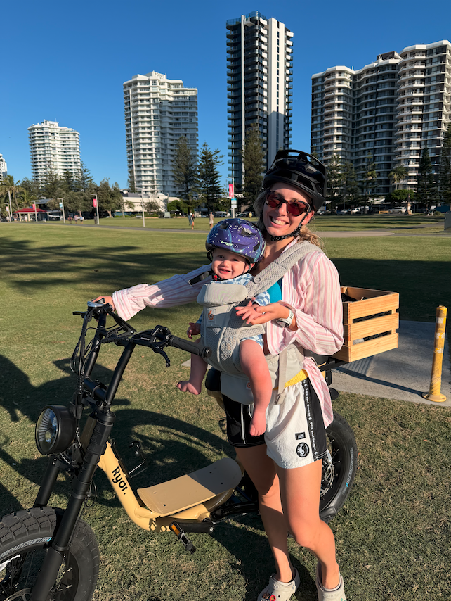 A woman wearing a helmet and sunglasses is smiling while holding a child in a baby carrier. The child is wearing a purple helmet and smiling. They are outdoors in a park with tall buildings in the background.