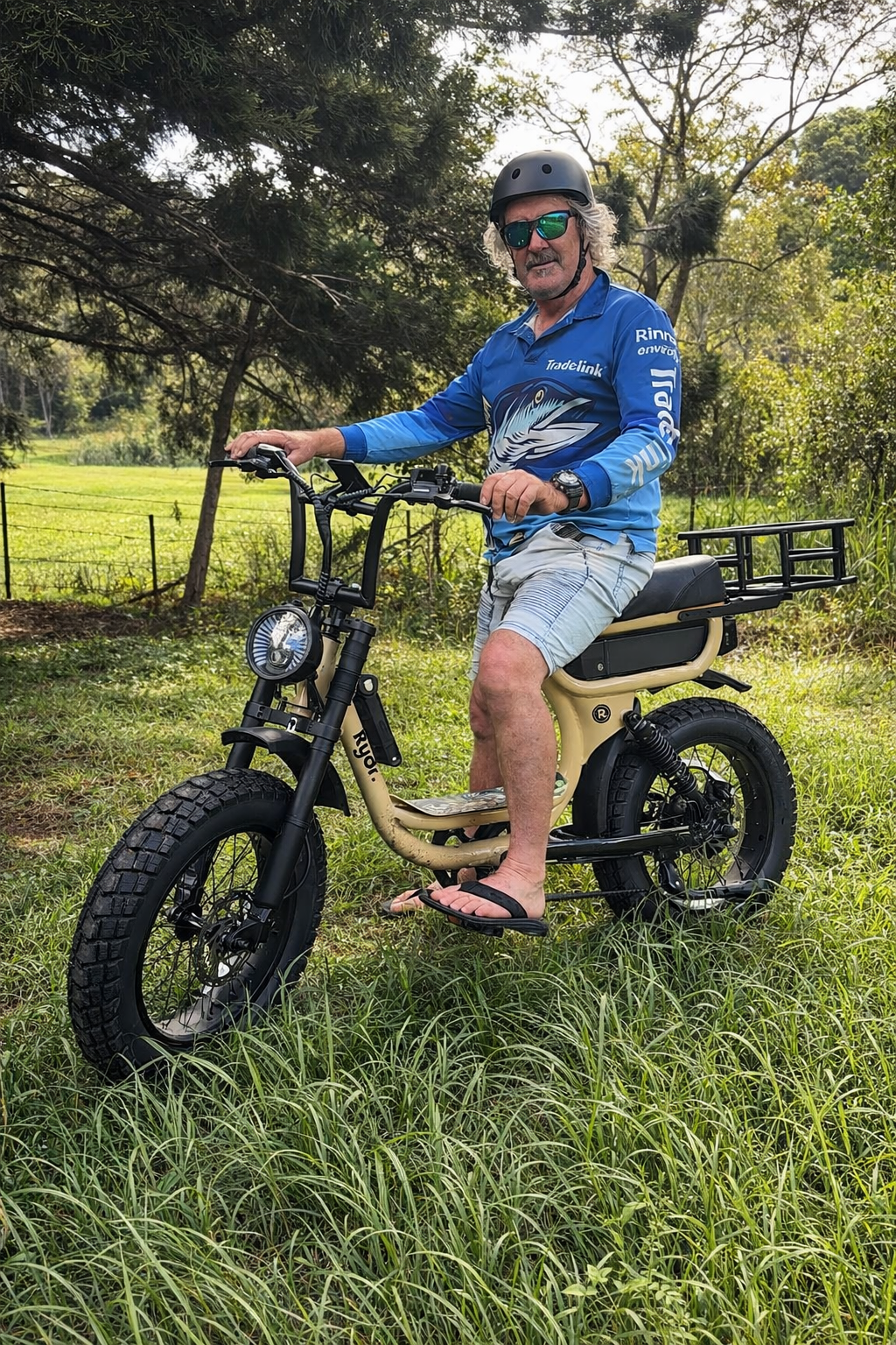 A man wearing a blue long-sleeve shirt, shorts, flip-flops, a helmet, and sunglasses is sitting on an electric bicycle in a grassy outdoor area with trees in the background.
