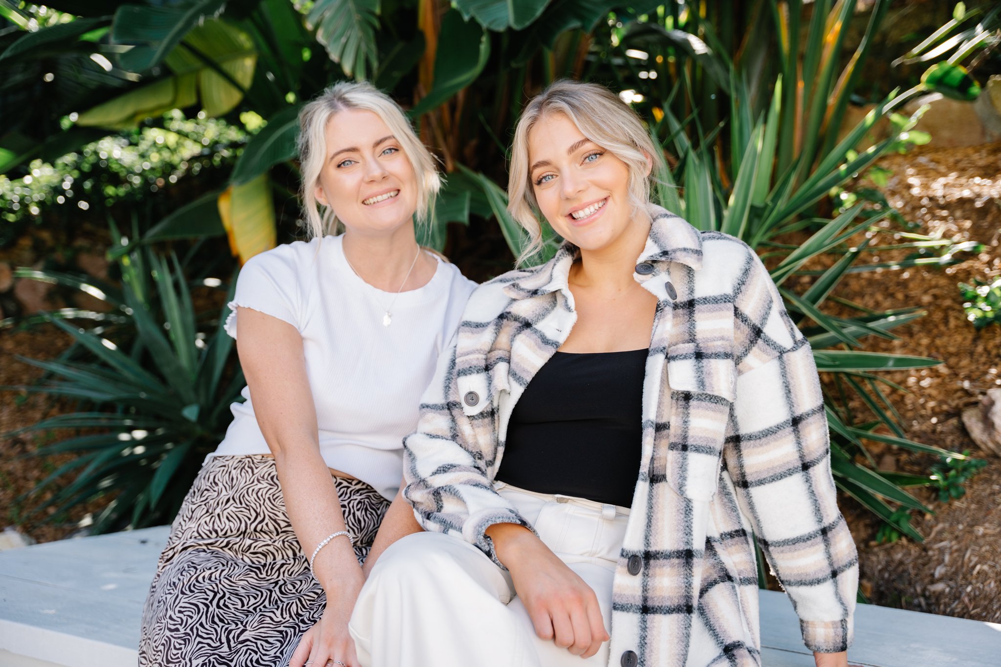 Two young women sitting outdoors in front of green plants, smiling at the camera. One wears a white shirt and a patterned skirt; the other wears a black top and a plaid shirt.