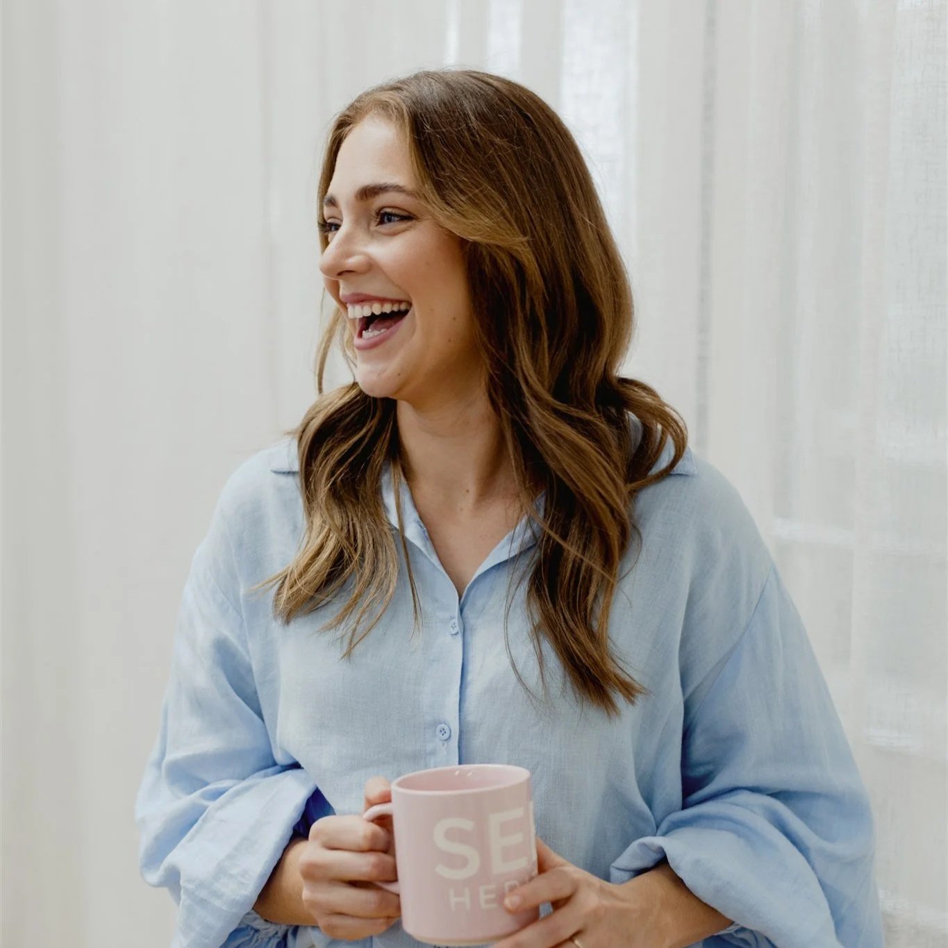 A woman with brown hair smiling and holding a pink mug indoors.