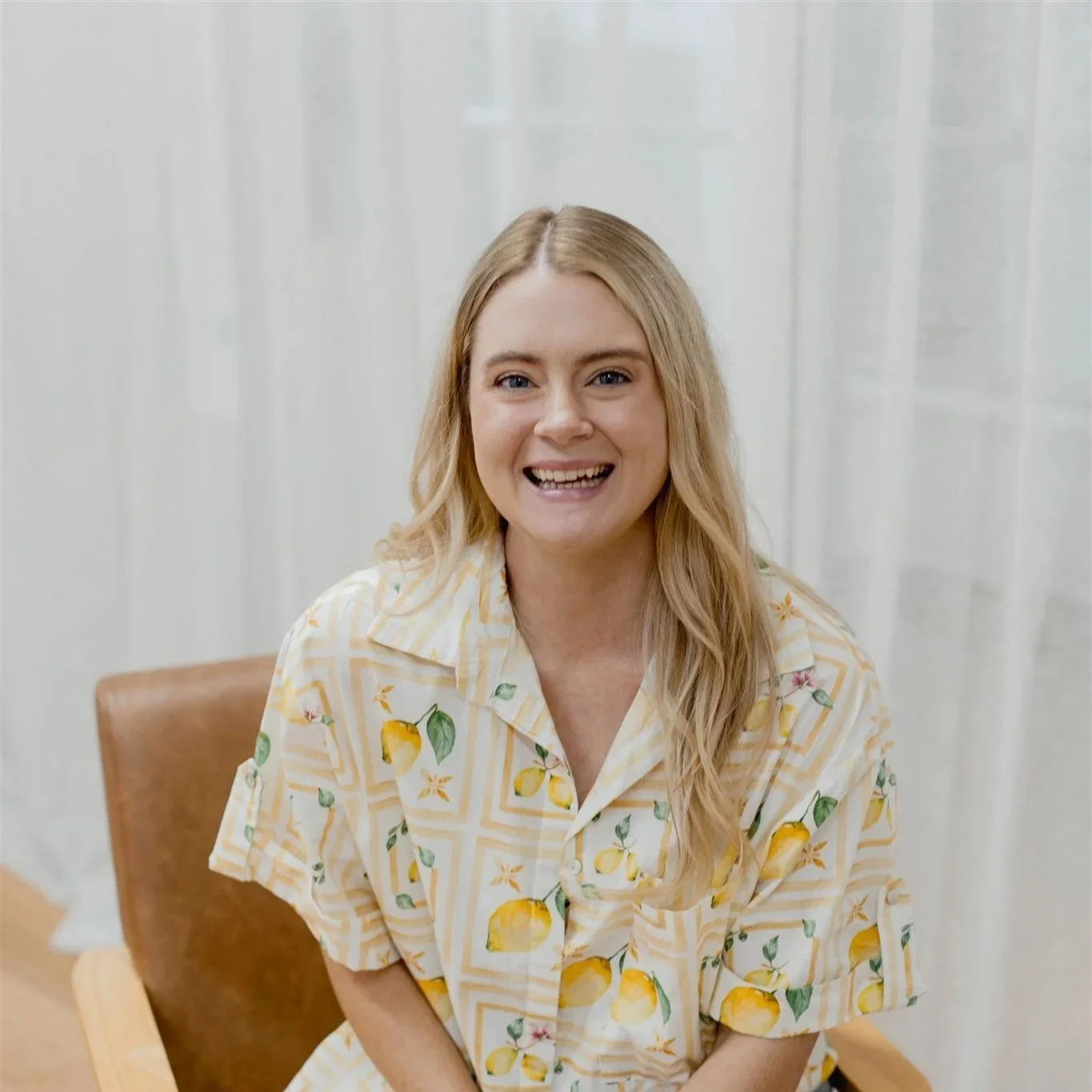 A smiling woman with blonde hair wearing a lemon-patterned shirt sitting on a wooden chair in front of a white curtain.