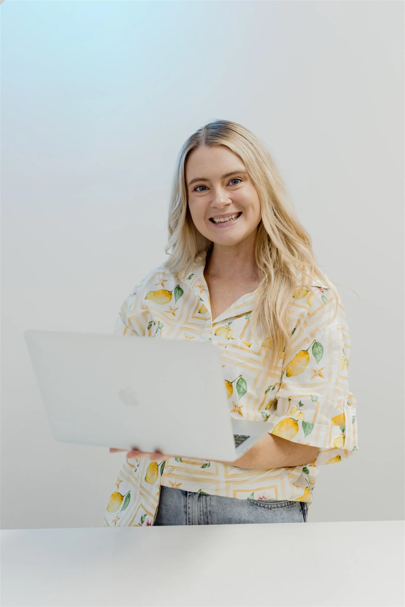 A woman with long blonde hair, smiling, holding a laptop, sitting at a white desk in a light-colored room.