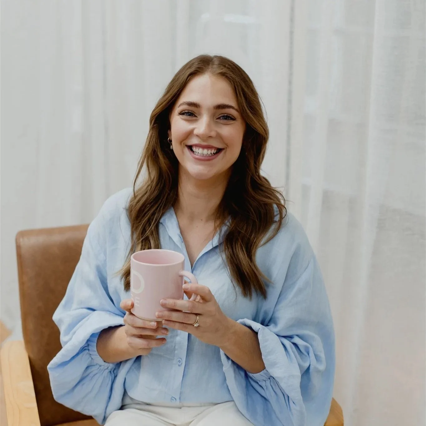 A smiling woman with long brown hair, wearing a light blue button-up shirt, holding a pink mug with both hands, sitting on a brown chair with a white curtain in the background.