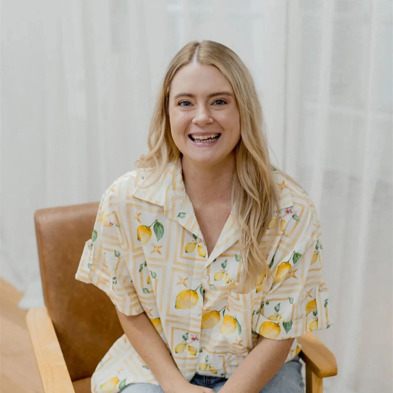 A woman with long blonde hair, smiling, wearing a short-sleeved button-up shirt with lemon pattern, sitting in a wooden chair with a brown cushion, in a room with light-colored curtains in the background.