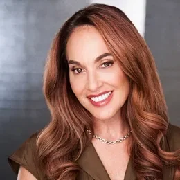 A woman with long, wavy auburn hair smiling at the camera, wearing a brown top and a pearl necklace.