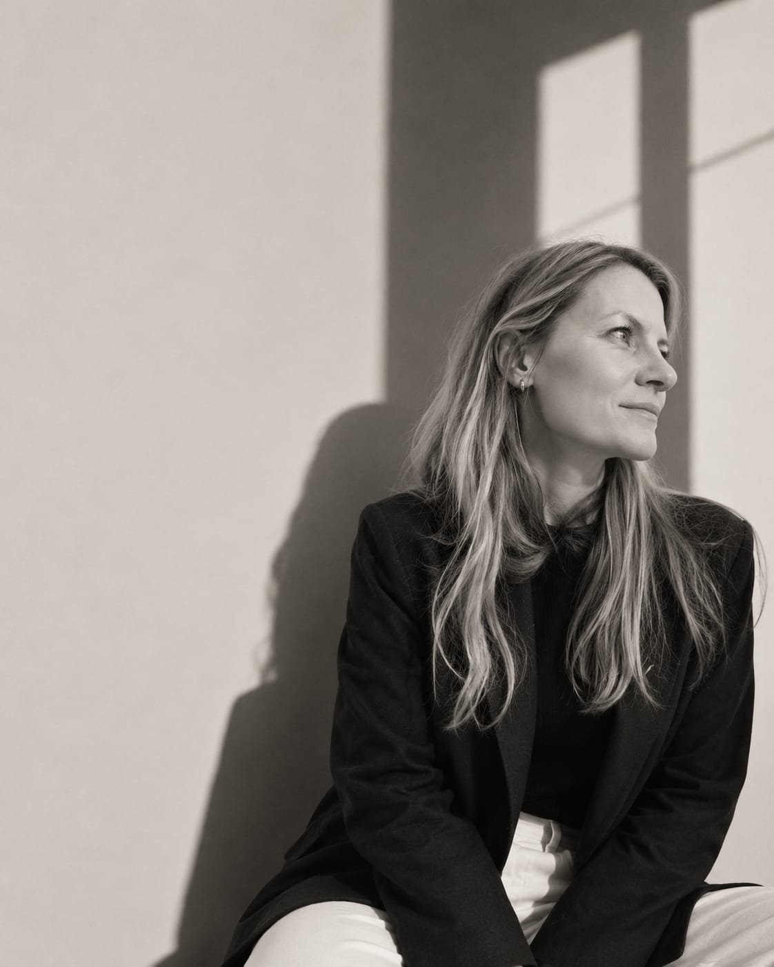 A black-and-white photo of a woman with long wavy hair, seated against a wall that casts a shadow and a window frame's shadow. She is wearing a dark blazer and light-colored pants, looking to the right with a faint smile.