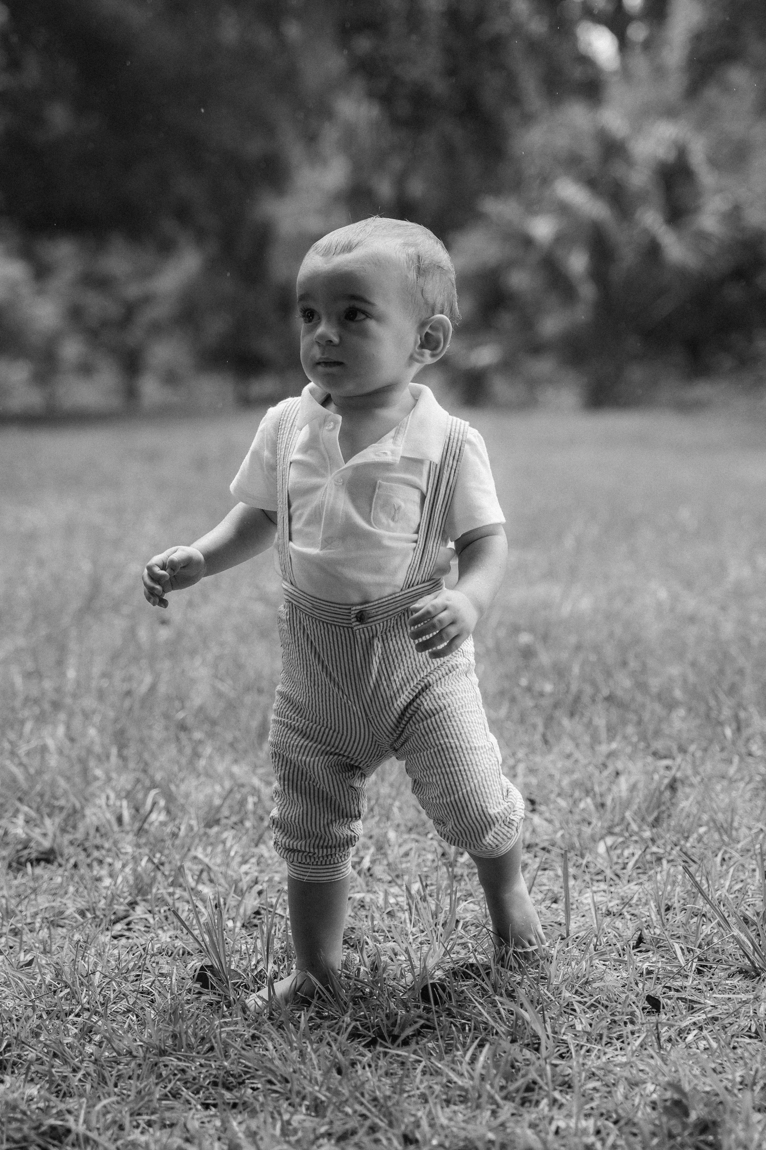 Honey+Haze Photography. A young child with short hair running outdoors on grass, wearing a short-sleeved shirt, suspenders, and pants, with a blurred background of trees.