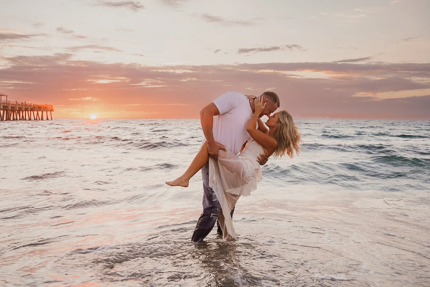 Honey+Haze Photography. A couple stands in shallow ocean water at sunset, the man lifting the woman in his arms while they kiss. The sky is painted with warm colors, and a pier extends into the water on the left side of the image.