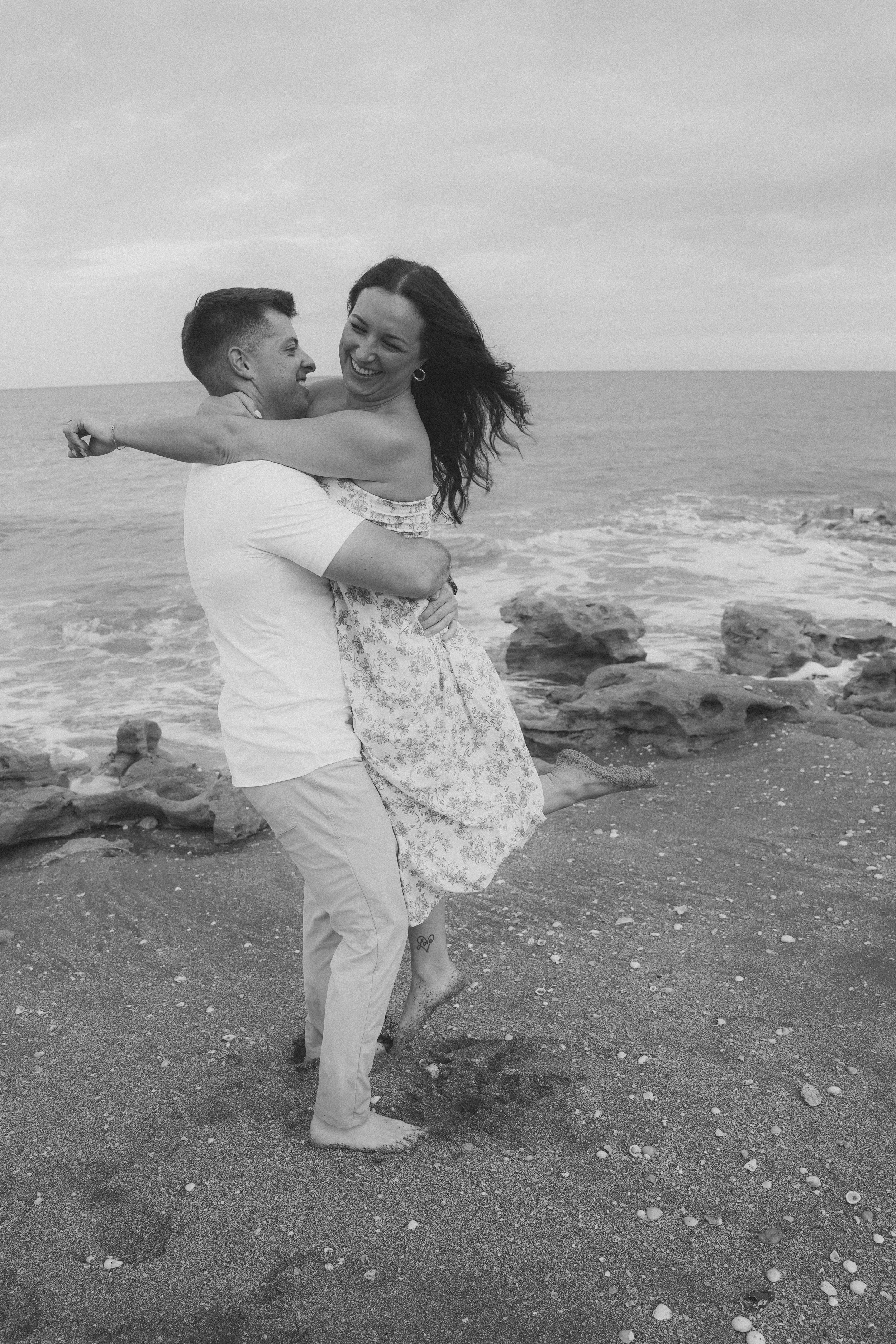 Honey+Haze Photography. A couple is embracing and laughing on a rocky beach with the ocean in the background. The man is lifting the woman, and both appear joyful. 