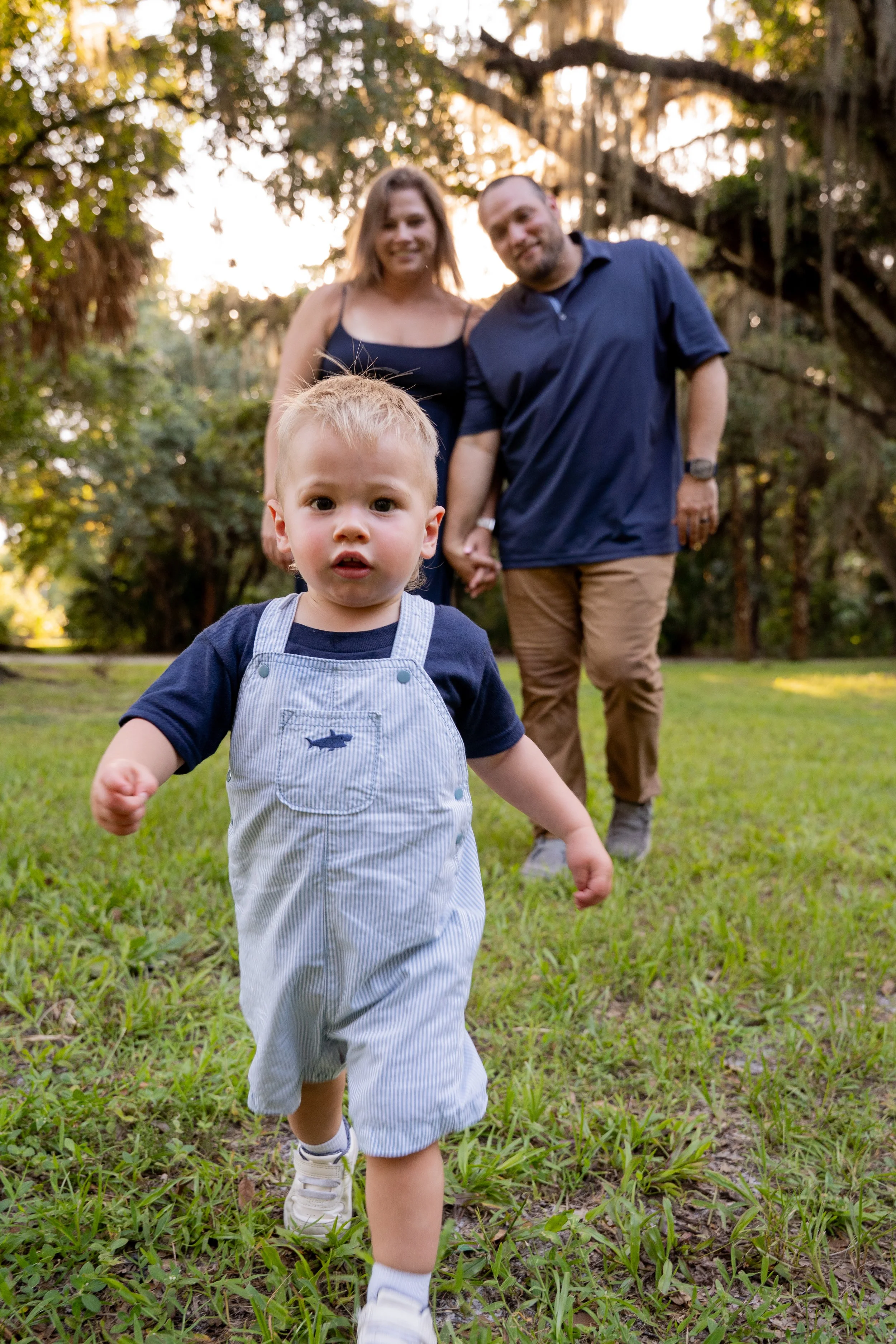 Honey+Haze Photography. A young boy in a blue shirt and striped overalls walking toward the camera on a grassy area with two adults holding hands behind him. The background features trees and sunlight filtering through the branches.