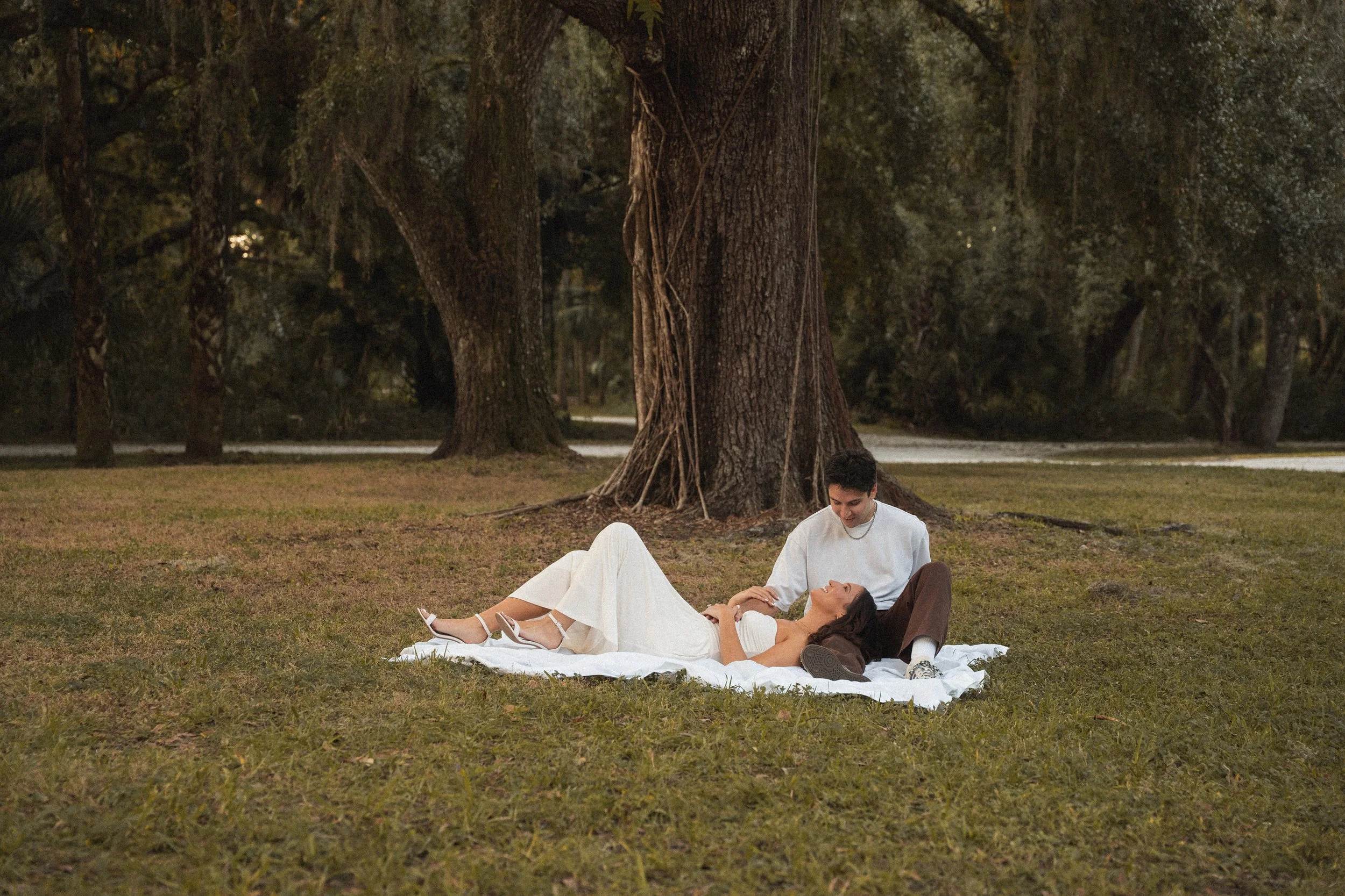 Honey+Haze Photography. A young couple is lying on a blanket under a large tree in a park The woman is lying down with her head resting on the blanket, while the man is sitting beside her, gently touching her arm.