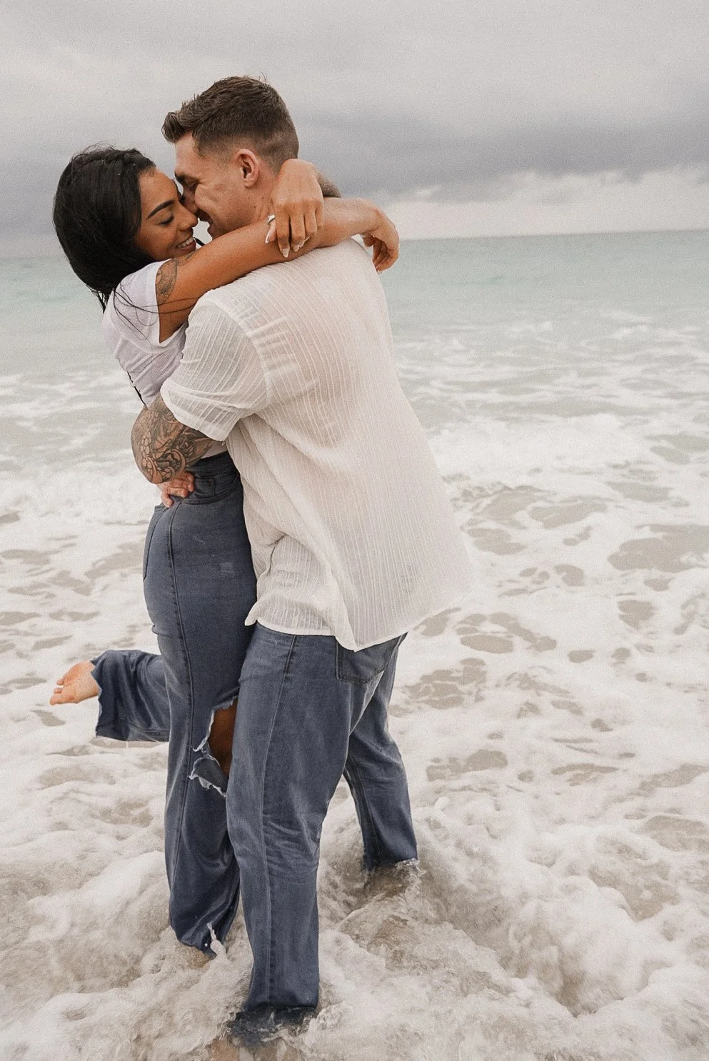 Honey+Haze Photography. A couple hugging and smiling on the beach, with cloudy sky and ocean waves in the background.
