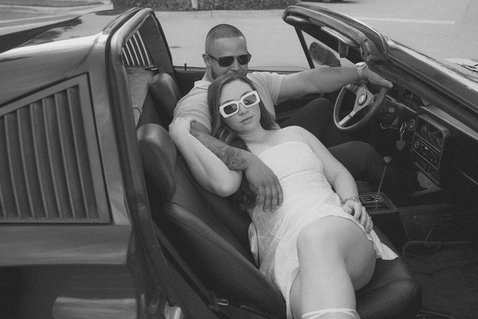 Honey+Haze Photography. A couple sitting in a convertible car parked outdoors, with the woman relaxing with her head on the man's lap and both wearing sunglasses.