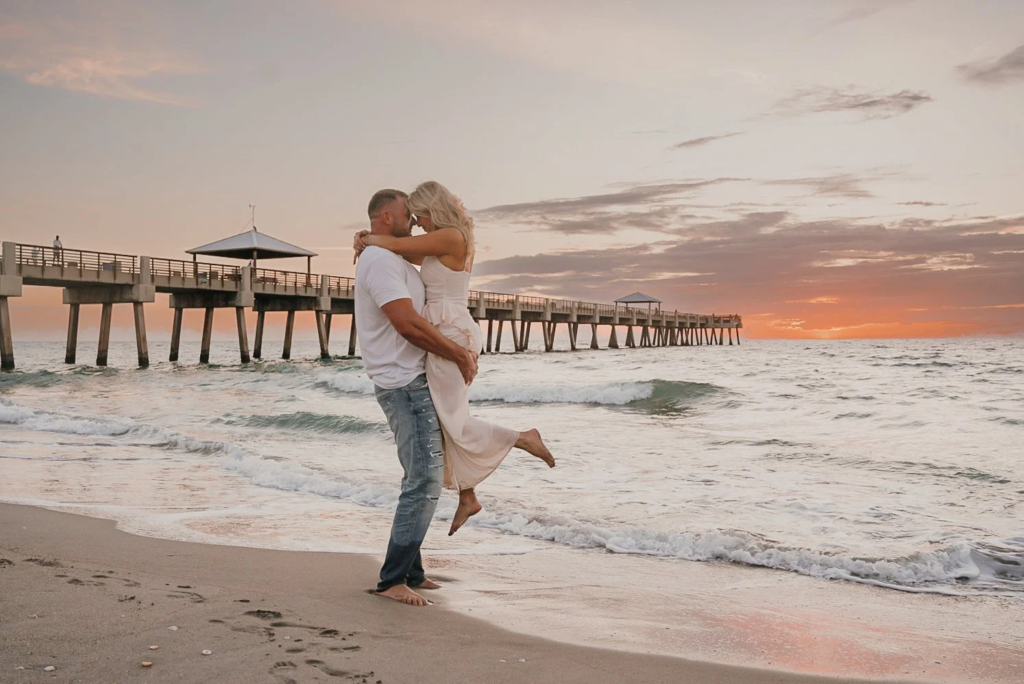 Honey+Haze Photography. A couple on the beach during sunset, with the man lifting the woman, near a pier extending into the water.