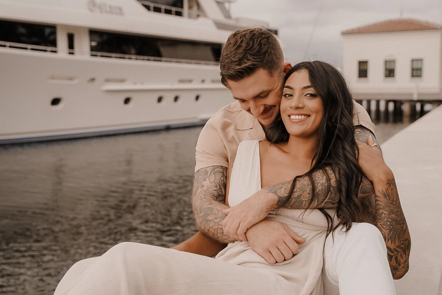 Honey+Haze Photography. A smiling couple sitting by the water in front of a yacht, with the man embracing the woman from behind.
