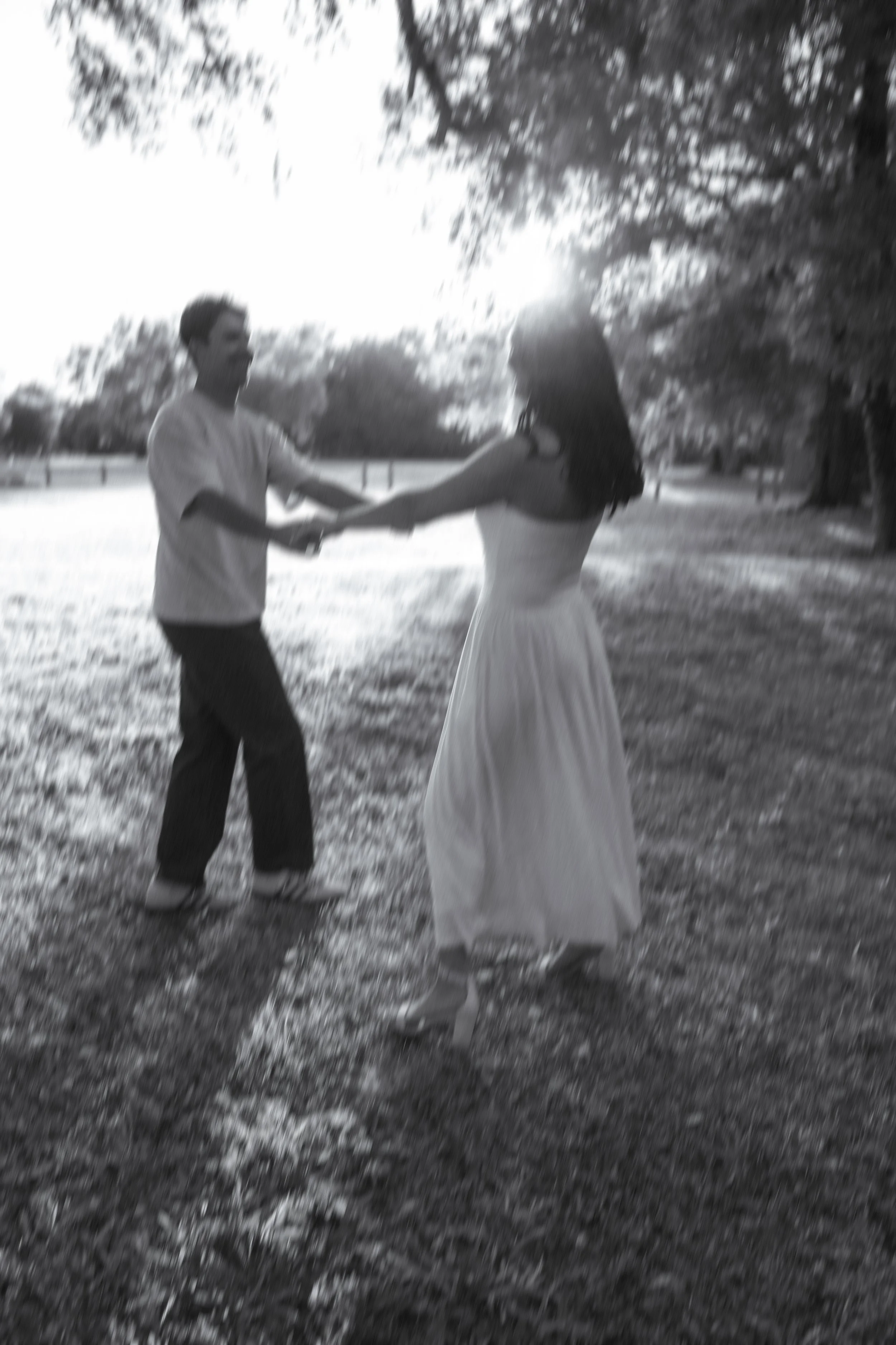 Honey+Haze Photography. A black-and-white photo of a man and woman dancing outdoors under a tree near a body of water. The man is holding the woman's hand, and both are smiling. The woman is wearing a long dress, and the man is dressed casually. 