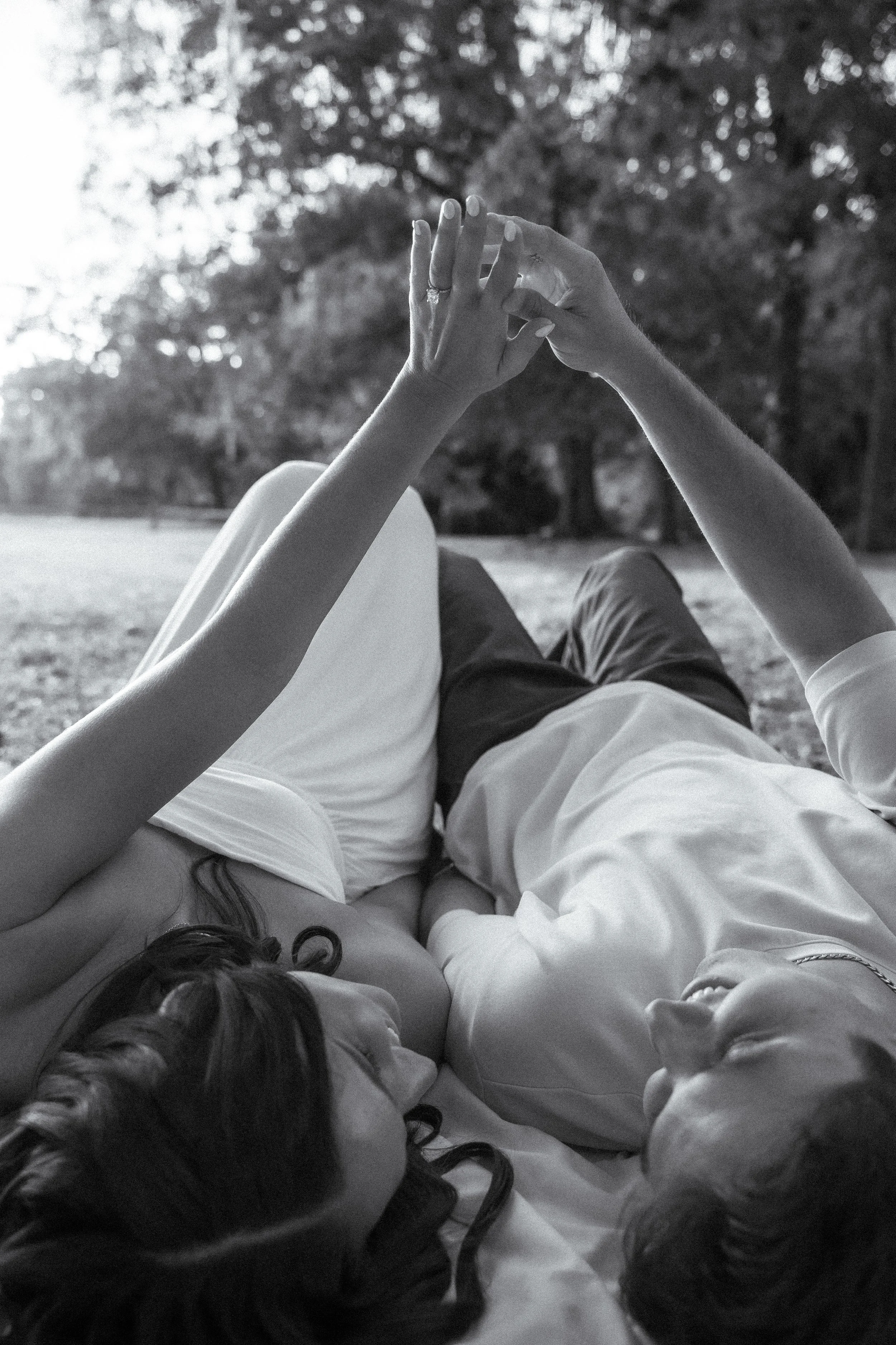 Honey+Haze Photography. A couple lying on grass outdoors, holding hands with rings visible, with trees in the background, black and white photograph.