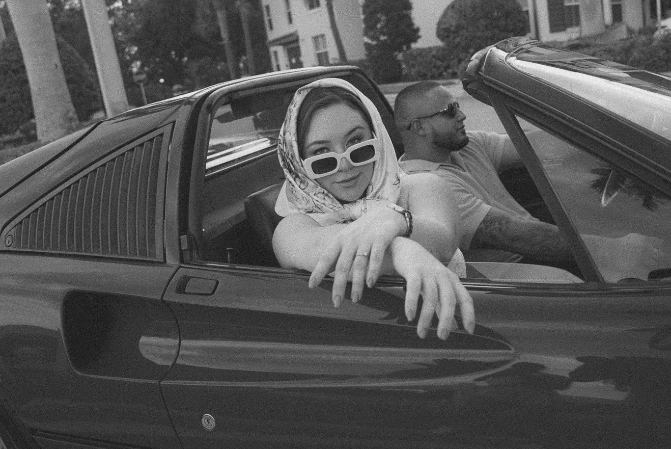 Honey+Haze Photography. A woman with sunglasses and a headscarf leaning out of a convertible car window, smiling, with a man driving in the background.