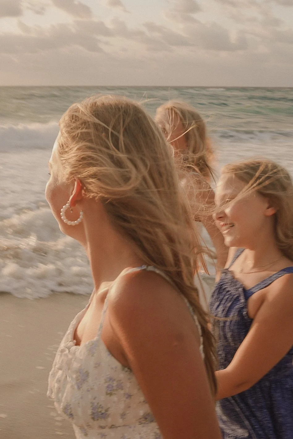 Honey+Haze Photography. Three girls with blonde hair enjoying a moment on the beach, with ocean waves in the background, during a cloudy day.